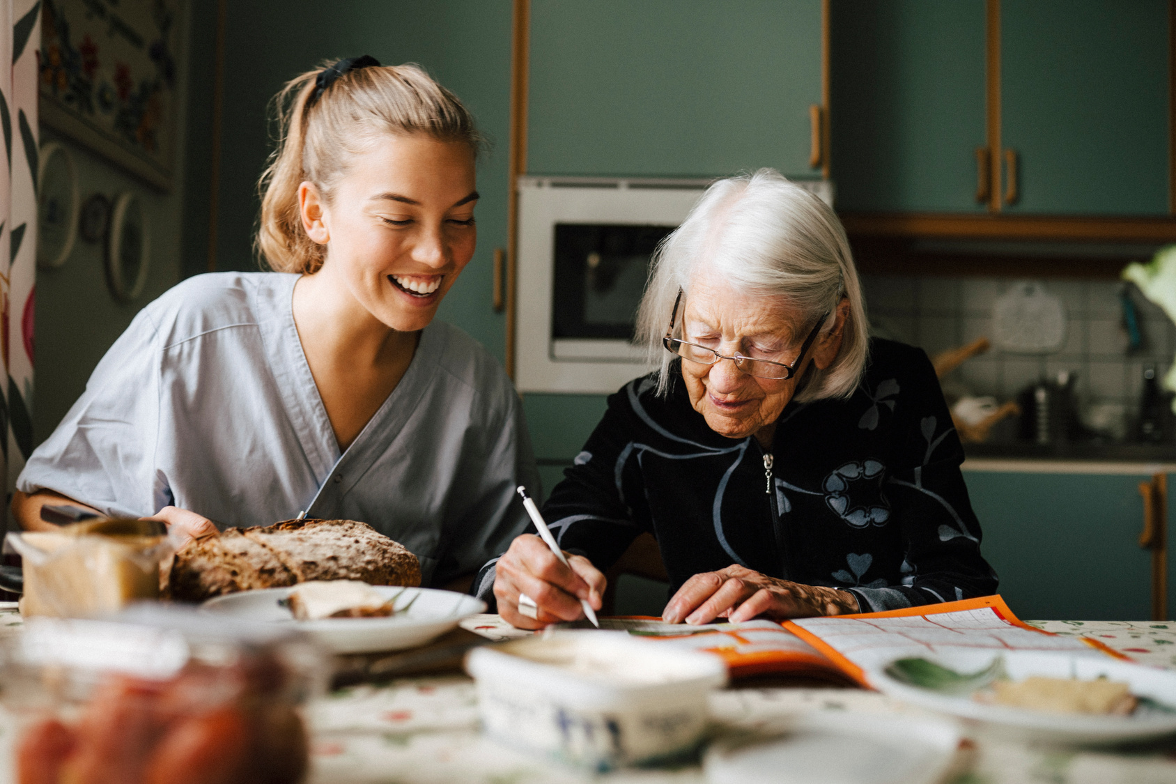 Seniorin und Pflegekraft sitzen am Tisch, beide Lächeln