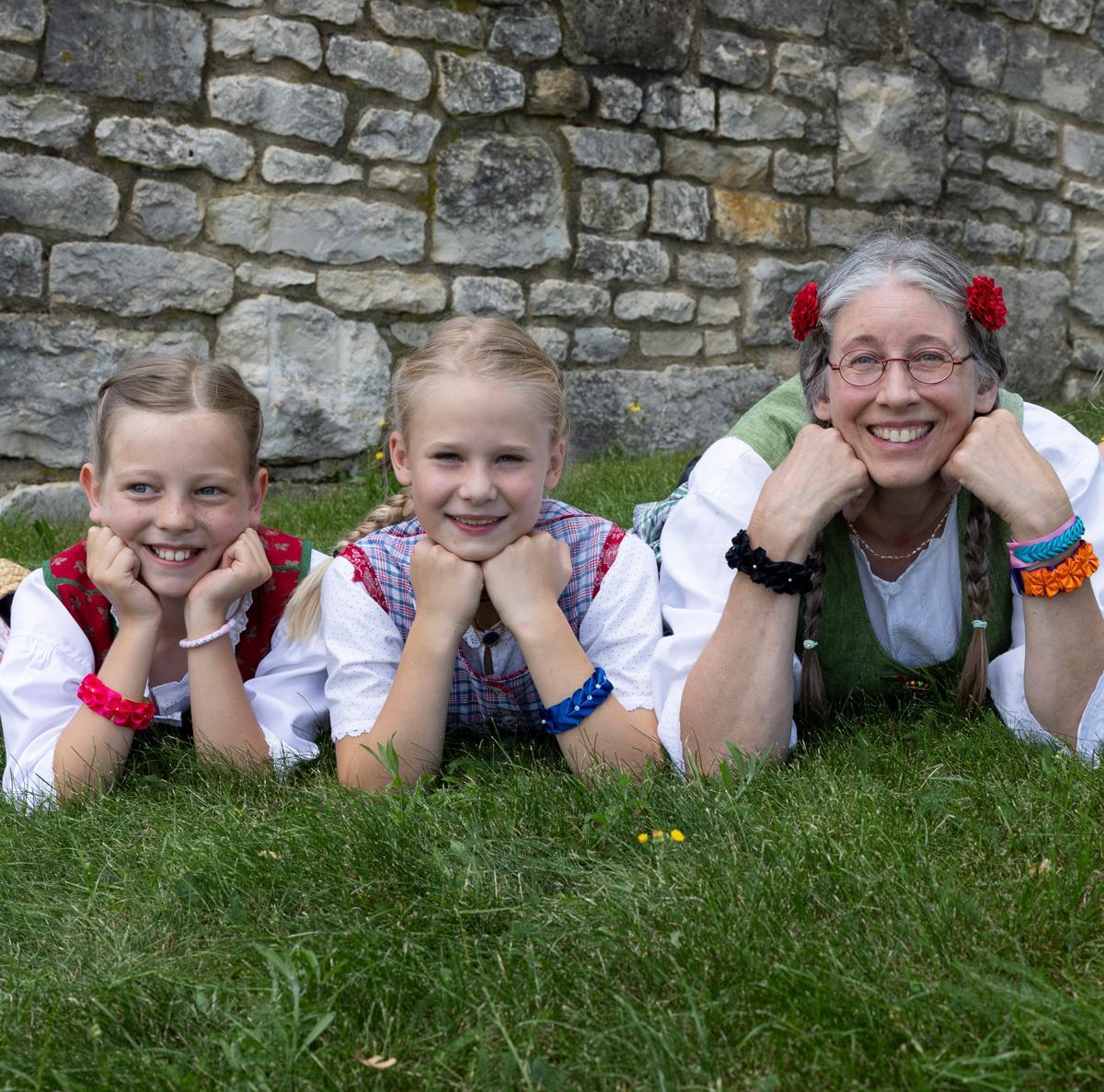 Zwei Mädchen und eine Frau liegen im Gras und schauen in die Kamera.