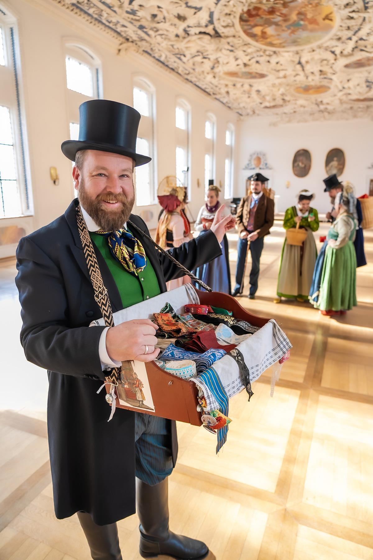 Ein Mann mit Bauchladen und eine Gruppe von Menschen in Tracht.