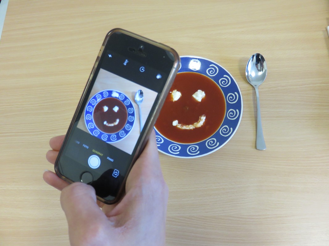 ein Teller Tomatensuppe, auf die mit Sahne ein Smiley gemalt ist, wird mit dem Smartphone fotografiert