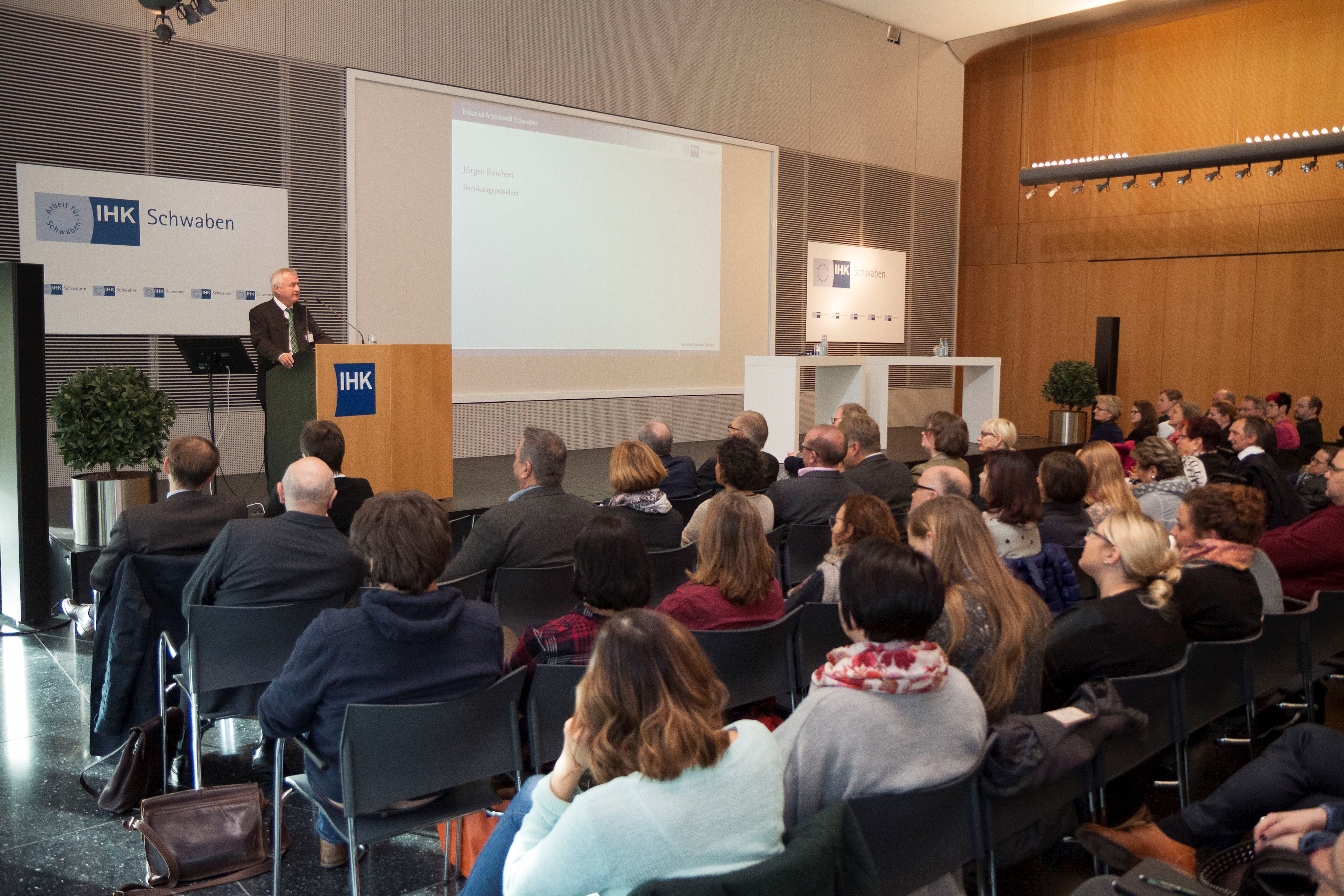 im Vordergrund das Publikum, im Hintergrund Bezirkstagspräsident Jürgen Reichert bei seinem Grußwort