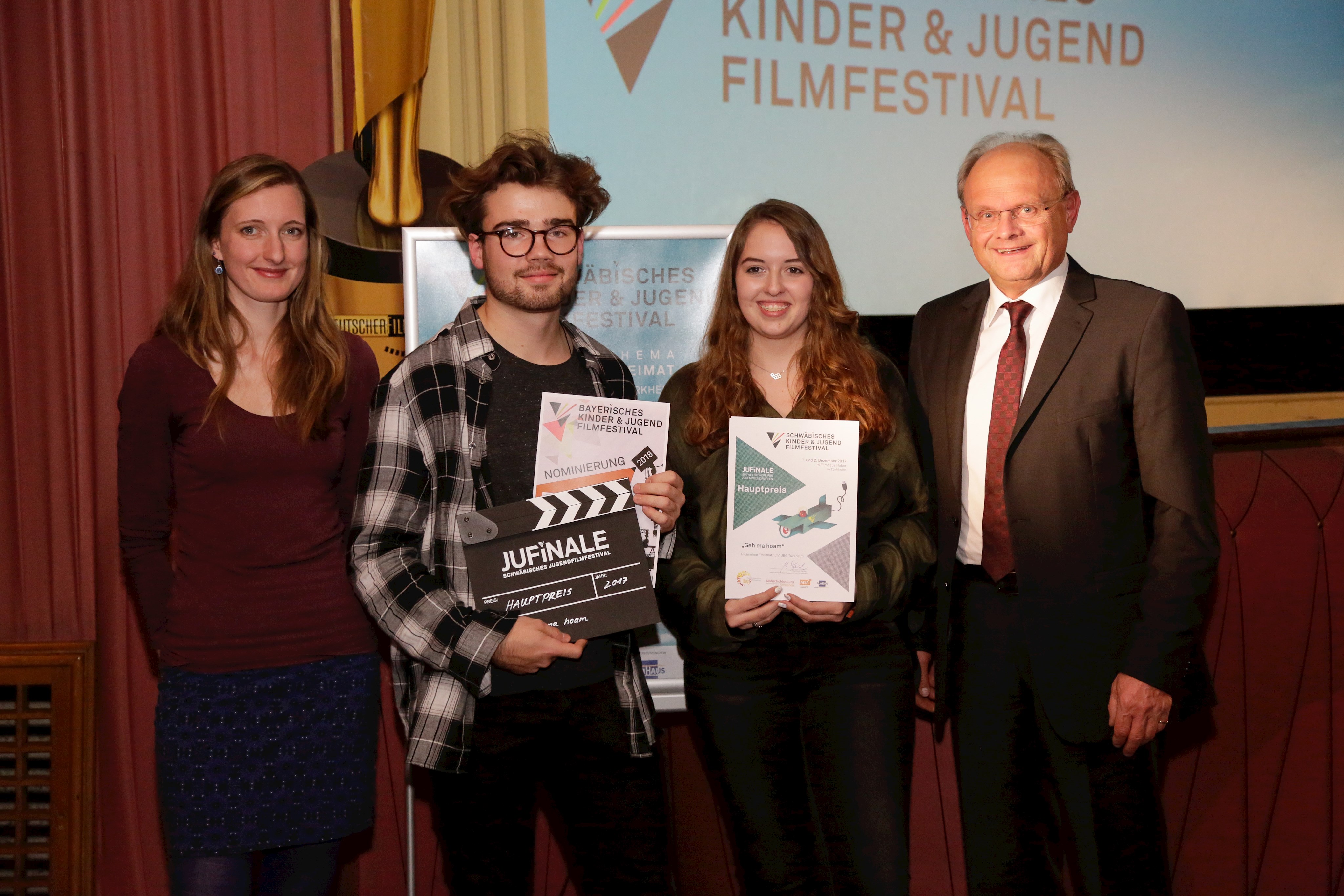 Gruppenbild vor der Kinoleinwand; die Preisträger halten Urkunden und den Preis (eine Filmklappe) in den Händen