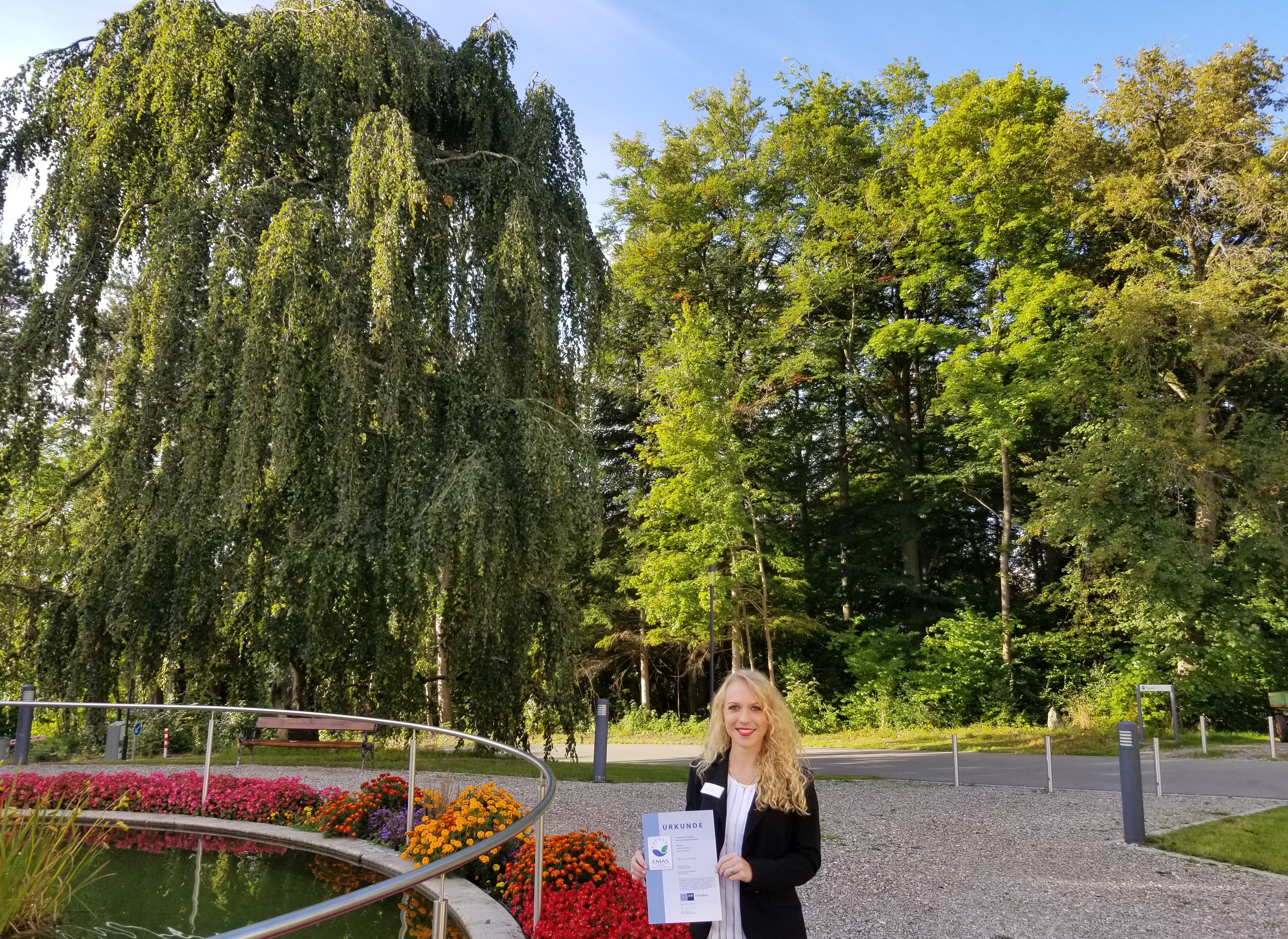 Manuela Kohler, Assistentin der Regionalleitung Süd und Umweltmanagementbeauftragte am BKH Kaufbeuren, freut sich inmitten von grünen Bäumen und bunten Blumen am Springbrunnen über die EMAS-Urkunde.