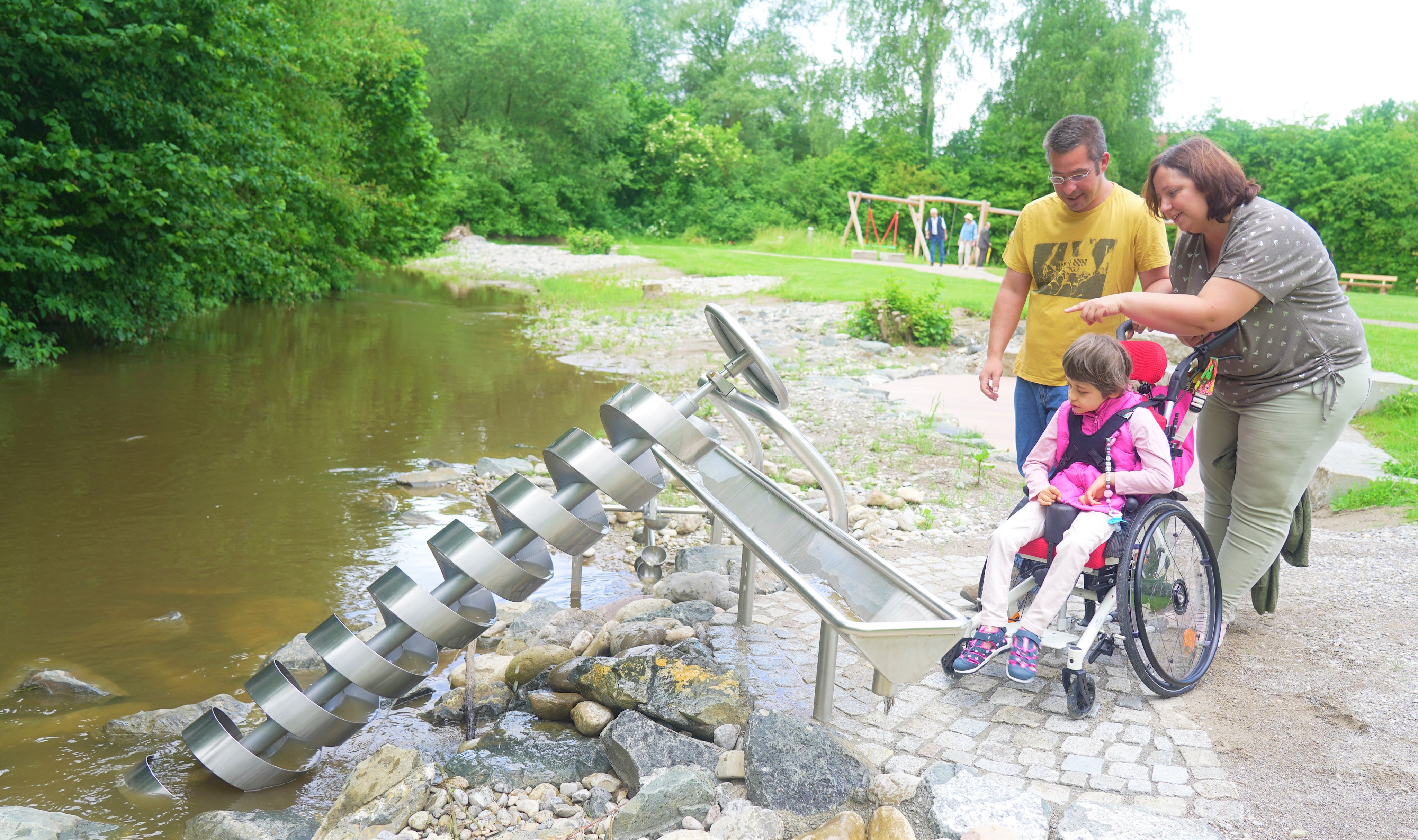 ein Mädchen im Rollstuhl und ihre Eltern an einem Wasserspielplatz